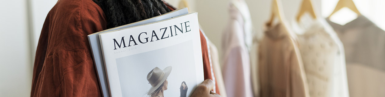 Eine Frau hält ein Modemagazin in der Hand. Im Hintergrund sind Kleider am Bügel zu erkennen.
