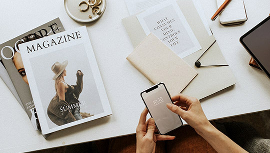 Magazine drucken Blick auf einen Schreibtisch mit gedruckten Magazinen, Notizbüchern und einer Person, die etwas am Smartphone eingibt